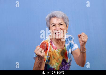 Une femme hispanique sénile attirante avec un grand sourire et des poings serrés se penchent vers l'avant dans l'excitation contre un fond bleu. Banque D'Images