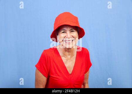 Belle femme hispanique senior souriante et vêtue d'une chemise rouge et d'un chapeau rouge isolé sur fond bleu. Banque D'Images