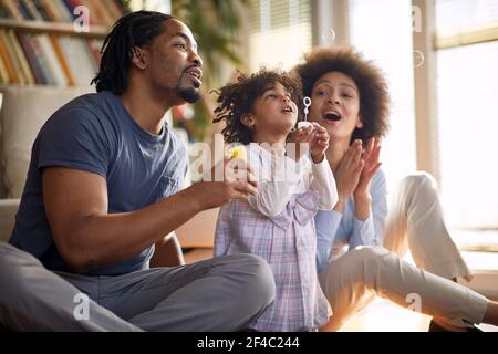 Les jeunes parents ont passé un bon moment avec leur petite fille mignonne dans une atmosphère gaie à la maison tout en faisant des bulles de savon ensemble. Famille, maison, pl Banque D'Images