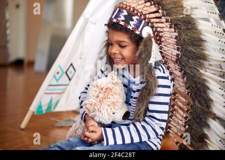 Un petit garçon mignon avec un capot de guerre indien tenant un fandy tout en étant assis sur le sol et jouant dans une atmosphère joyeuse à la maison. Famille, maison, Playti Banque D'Images