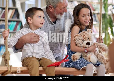 Les petits-enfants sont assis sur la balançoire et jouent avec leur grand-père dans une atmosphère ludique à la maison. Famille, maison, temps de jeu Banque D'Images