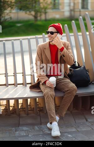 Homme hipster souriant dans des lunettes de soleil parlant sur le téléphone portable, assis sur le banc tenant une tasse de papier de café Banque D'Images