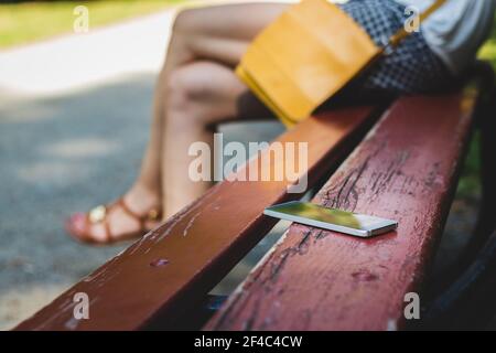 Femme assise dans un parc public et oubliée le smartphone sur le banc. Téléphone mobile perdu. Banque D'Images