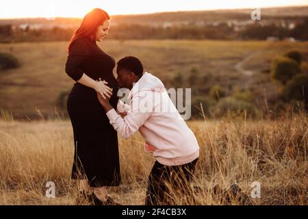 Couple de mariage heureux sur le terrain, mari surjoyeux baiser ventre enceinte de belle femme, jeune famille s'attendent à un bébé, excité par la parentalité Banque D'Images