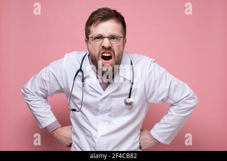 Un médecin mécontent portant des lunettes, une blouse blanche et un stéthoscope au niveau du cou hurlent avec colère tout en regardant dans l'appareil photo. Banque D'Images