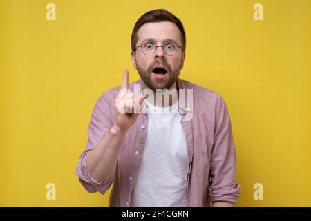 Bonne idée. Le beau homme en lunettes montre l'index vers le haut, regarde la caméra avec sa bouche ouverte et a un visage intrigant. Banque D'Images
