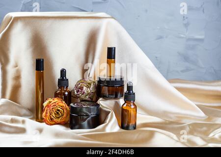 Divers contenants pour le stockage de cosmétiques en verre foncé se trouvent sur les plis de tissu satiné de soie, entouré de fleurs de rose séchées. Parfum, sérum ou Banque D'Images