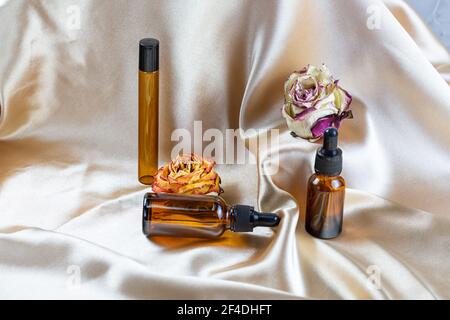 Divers contenants pour le stockage de cosmétiques en verre foncé se trouvent sur les plis de tissu satiné de soie, entouré de fleurs de rose séchées. Parfum, sérum ou Banque D'Images