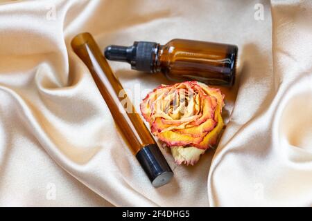 Divers contenants pour le stockage de cosmétiques en verre foncé se trouvent sur les plis de tissu satiné de soie, entouré de fleurs de rose séchées. Parfum parfum c Banque D'Images