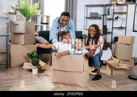 Joyeux parents africains et deux enfants jouant avec des boîtes pendant la réinstallation. De jolies filles vous aident à déballer vos boîtes avec plaisir. Des moments heureux de journée mouvante. Banque D'Images