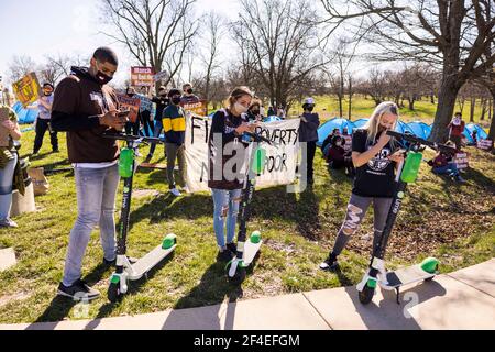 Un groupe de fans de basket-ball se connecte à l'application Lime Scooter tandis que les manifestants derrière eux protestent en faveur des membres de la communauté Bloomington sans hébergement pendant le tournoi NCAA. Une coalition de membres de la communauté Bloomington, des activistes, Et les étudiants de l'Université de l'Indiana défilent de Dunn Meadow à la salle d'assemblée Simon Skjodt lors de la « Marche pour mettre fin à la folie » pour protester contre le traitement par la ville de Bloomington des résidents sans logement qui latent des tentes sur des biens publics et le paiement de départ de 10 millions de dollars que l'Université de l'Indiana devra payer Archie Miller, entraîneur de basket-ball pour hommes. Plusieurs jeux Banque D'Images