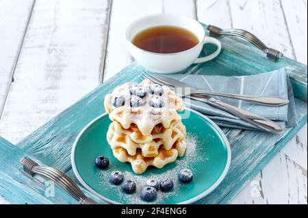 Gaufres maison à la vanille avec poudre de sucre et bleuets de fougères sur une assiette, petit déjeuner familial parfait. Banque D'Images