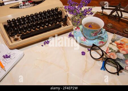 Machine à écrire manuelle rétro à l'ancienne sur un bureau avec tasse, bloc-notes, lunettes, pèse-lettres et fleurs avec espace de copie. Banque D'Images