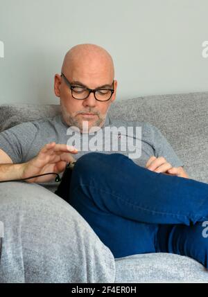 Chauve a dirigé un homme d'âge moyen assis sur un canapé et regardant vers le bas Comme il utilise son iPad Banque D'Images