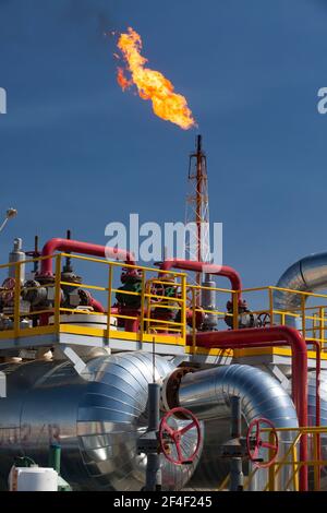 Usine pétrochimique (raffinerie de pétrole). Tuyaux, tubes, vannes et échangeur de chaleur sur torche à gaz orange géant et tour sur fond bleu ciel. Banque D'Images