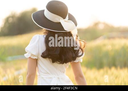 Vue arrière d'une jeune femme portant un chapeau noir et une robe blanche marchant seule sur un champ d'orge en fin d'après-midi avant le coucher du soleil, lumière chaude d'heure dorée Banque D'Images