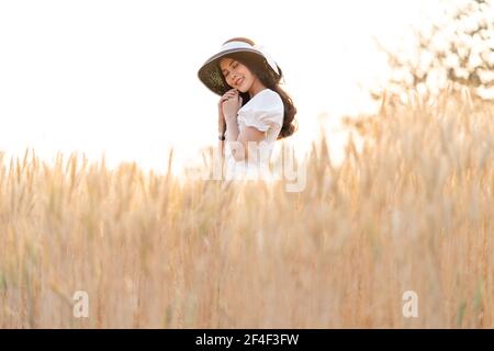 Bonne jeune belle femme portant un chapeau noir et une robe blanche en s'appréciant rêvant de jour dans l'orge d'or classé en fin d'après-midi, rétro-éclairé, b Banque D'Images