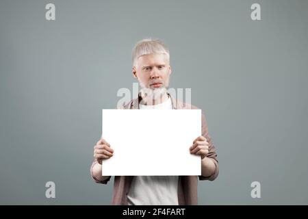 Albino homme tenant une feuille de papier vide avec l'espace libre, posant à l'appareil photo sur fond gris, publicité et maquette Banque D'Images