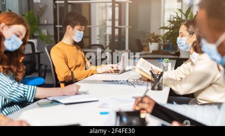 Des gens internationaux portant des masques chirurgicaux en utilisant un ordinateur portable assis au bureau Banque D'Images