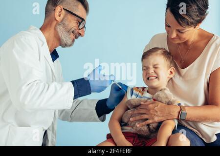 Médecin injectant sur le bras d'un garçon en fermant les yeux avec douleur. Un professionnel de la santé de sexe masculin donne le vaccin à un enfant assis avec sa mère. Banque D'Images