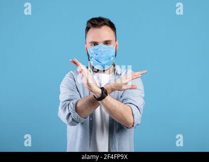 Lutte contre le coronavirus. Jeune homme caucasien dans le masque de visage gesturant ARRÊTER sur fond bleu studio Banque D'Images