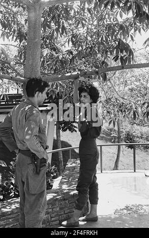 Photo de profil pleine longueur de Celia Sanchez Manduley, debout sous un arbre avec sa main accrochée autour d'une branche basse, parlant avec un administrateur armé et en uniforme au parc national de la Guira, Pinar del Rio, Cuba, 1964. De la collection de photographies Deena Stryker. () Banque D'Images