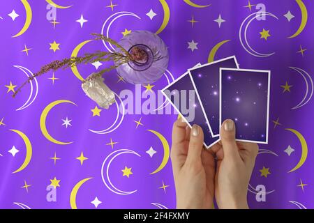 Mains de jeune femme avec vernis à ongles doré tenant trois cartes de tarot ou d'oracle, sur une étoile violette et un tissu de table à motif croissant Banque D'Images