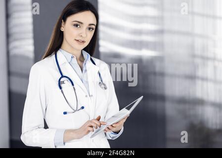 Gaie et intelligente femme-médecin tient une tablette dans ses mains, alors qu'elle est debout dans une clinique. Portrait d'une femme médecin sympathique Banque D'Images