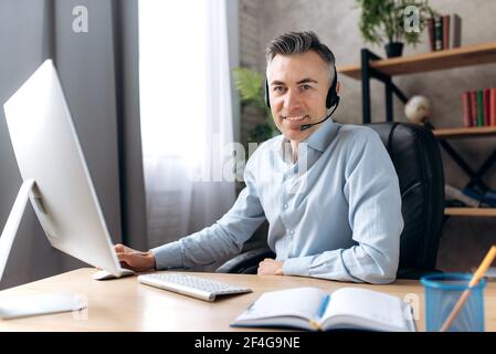 Portrait d'un homme caucasien adulte confiant et réussi en tenue officielle, d'un homme d'affaires ou d'un avocat assis sur le lieu de travail dans un bureau moderne à l'aide d'un ordinateur et d'un casque, en regardant l'appareil photo avec un sourire amical Banque D'Images