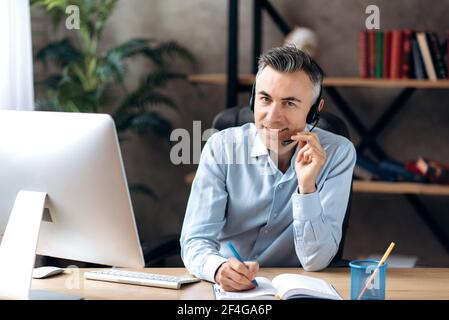 Un homme d'affaires intelligent et réussi s'assoit à la tenue et au micro-casque modernes de bureau, communique avec un client ou des collègues prend des notes dans un ordinateur portable, regarde l'appareil photo et sourit Banque D'Images