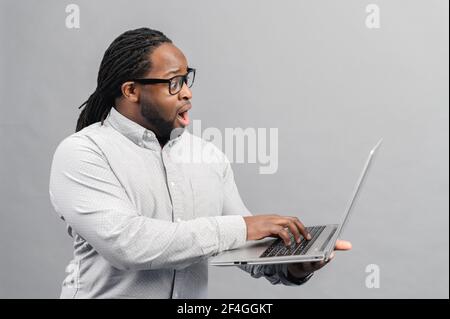 Homme noir debout avec ordinateur portable sur fond gris, dactylographie, concept de l'homme émotions, se sentant stupéfait par les nouvelles en ligne regardant l'écran d'ordinateur, investisseur de Trader surpris par les changements de la bourse Banque D'Images