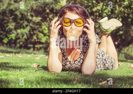 Bonne fille avec un casque écouter de la musique et gonfle une gomme à mâcher. Jeune femme élégante dans un style décontracté et lunettes de soleil jaunes à l'extérieur Banque D'Images