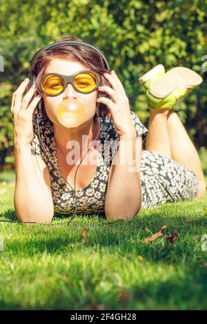 Bonne fille avec un casque écouter de la musique et gonfle une gomme à mâcher. Jeune femme élégante dans un style décontracté et lunettes de soleil jaunes à l'extérieur Banque D'Images