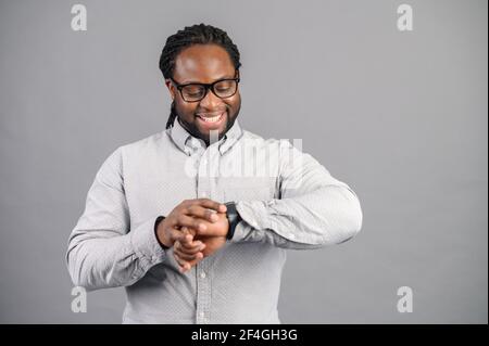 Homme d'affaires afro-américain ponctuel de bonne humeur, debout, vérifiant sa montre intelligente, reçu une notification, vérifie le pouls à l'aide d'un appareil moderne, suit l'heure lors d'une réunion ou d'une date Banque D'Images