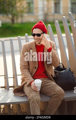 Homme hipster souriant dans des lunettes de soleil parlant sur le téléphone portable, assis sur le banc tenant une tasse de papier de café Banque D'Images