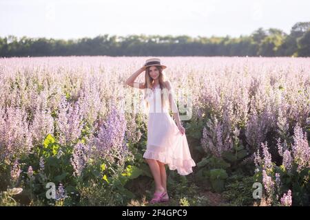 Jeune femme blonde en mousseline légère sur fond de champ de fleur de sauge rose. Portrait de belle fille tenant chapeau de paille. Semaine Banque D'Images