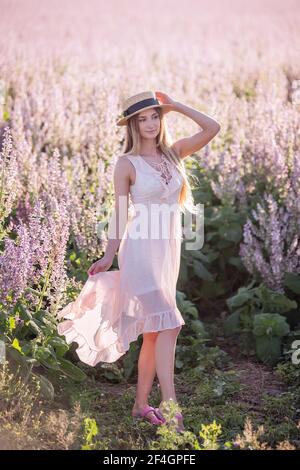 Jeune femme blonde en mousseline légère sur fond de champ de fleur de sauge rose. Portrait de belle fille tenant chapeau de paille. Semaine Banque D'Images