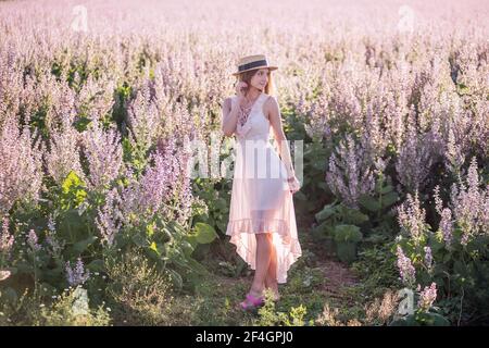 Jeune femme blonde en mousseline légère sur fond de champ de fleur de sauge rose. Portrait de belle fille tenant chapeau de paille. Semaine Banque D'Images