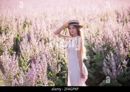 Jeune femme blonde en mousseline légère sur fond de champ de fleur de sauge rose. Portrait de belle fille tenant chapeau de paille. Semaine Banque D'Images