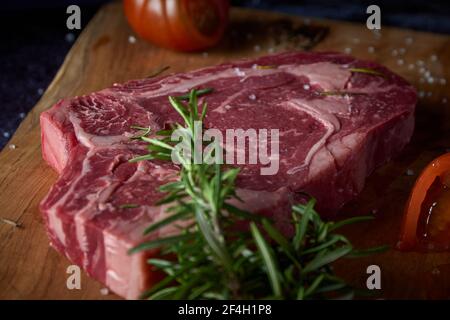 Steak de bœuf frais et cru sur une planche de bois avec sel et autres assaisonnements sur une surface sombre. Steak prêt à griller. Nourriture sombre. Viande, tomate, poivre, citron Banque D'Images