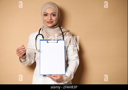 Charmante femme arabe musulmane médecin dans hijab pointant avec un crayon sur une feuille de papier vierge sur un bloc-notes Santé et concept d'assurance. Station santé Copy Banque D'Images