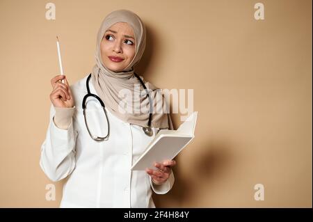 Une femme arabe musulmane, médecin attirante, tient un journal intime et regarde avec soin, pointant avec un crayon blanc sur un fond beige avec un espace de copie. Banque D'Images