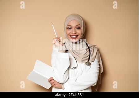 Jolie femme arabe musulmane médecin tenant un journal et regardant l'appareil photo avec un beau sourire en dents de souris, pointant avec un crayon blanc sur un b beige Banque D'Images