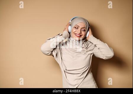 Une femme en hijab et casque sourit et se détend tout en écoutant la musique sur fond beige avec un espace de copie. Émotion. Banque D'Images
