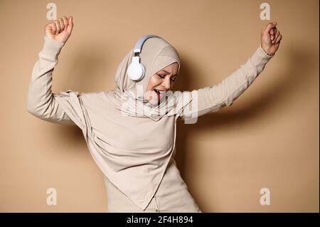Une femme musulmane heureuse dans le hijab et le casque sourit et se détend, danse tout en écoutant la musique sur fond beige avec l'espace de copie Banque D'Images