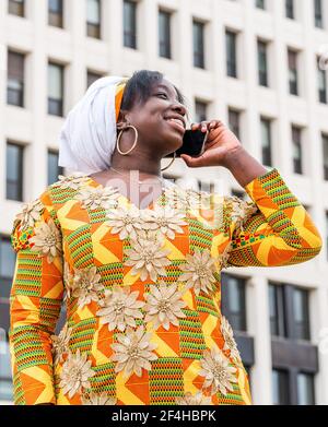 De dessous de la femme noire souriante dans les vêtements d'ornement parlant sur un téléphone portable tout en regardant loin dans la ville Banque D'Images