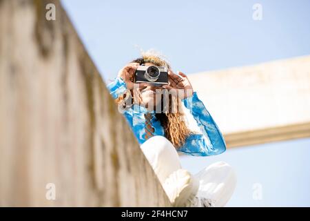 D'en-dessous de la femme ethnique aux cheveux mauris heureux dans élégant tenue assise sur une clôture en béton et prise de photos rétro appareil photo en été ensoleillé Banque D'Images