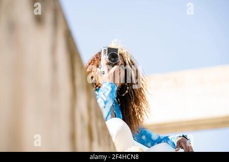 D'en-dessous de la femme ethnique aux cheveux mauris heureux dans élégant tenue assise sur une clôture en béton et prise de photos rétro appareil photo en été ensoleillé Banque D'Images