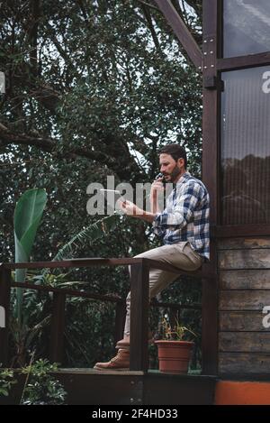 Vue latérale d'un architecte homme sérieux assis sur une rambarde terrasse en bois et parler sur un smartphone tout en discutant du projet Banque D'Images