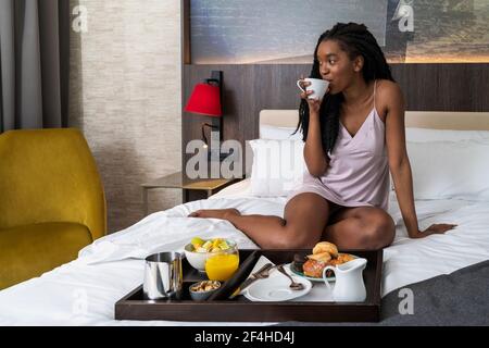 Tout le contenu jeune afro-américaine touristique dans vêtements de nuit sur un lit confortable avec plateau de délicieux petit déjeuner et boire du café Banque D'Images